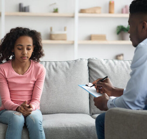 Upset african american girl having conversation with child psychologist Upset african american curly girl sitting on couch at clinic, having conversation with child psychologist black man, answering questions and looking down. Behavioral disorders in teenagers